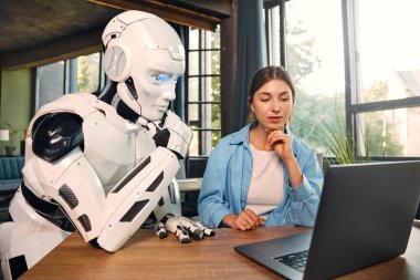 Genç bir kadın ve insansı bir robot modern bir ofiste dizüstü bilgisayarın başında otururken çalışıyorlar. İnsanlar ve yapay zeka arasındaki işbirliği.