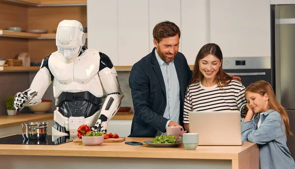 Robot housekeeper preparing food in the kitchen while family with daughter using laptop. Living together between humans and artificial intelligence.