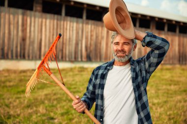 Çiftlikteki bir ahırın arka planında dirgenle duran olgun, yakışıklı bir erkek çiftçi. Çiftçilik ve tarım kavramı.