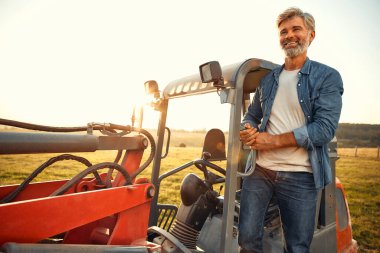 Olgun bir erkek çiftçi sıcak bir günde bir tarlada bir traktörde oturup çiftlikte çalışıyor. Çiftçilik ve tarım kavramı.