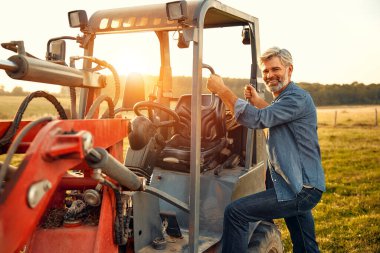 Olgun bir erkek çiftçi sıcak bir günde bir tarlada bir traktörde oturup çiftlikte çalışıyor. Çiftçilik ve tarım kavramı.