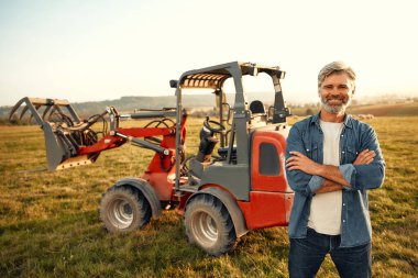Olgun bir erkek çiftçi sıcak bir günde bir tarlada traktörün yanında duruyor ve çiftlikte çalışıyor. Tarım ve tarım kavramı.