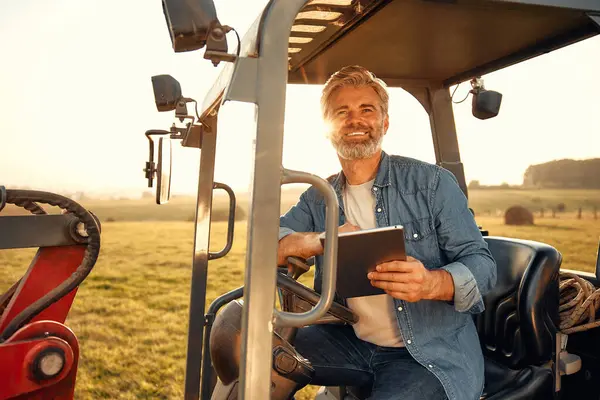 Mature handsome male farmer with a tablet sitting in a tractor on a field on a hot day working on a farm. Farming and agriculture concept.
