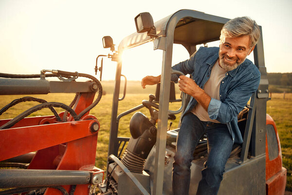 Mature handsome male farmer sitting in a tractor on a field on a hot day working on a farm. Farming and agriculture concept.
