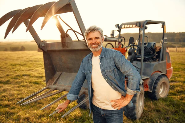 Mature handsome male farmer standing near a tractor on a field on a hot day and working on the farm. Agriculture and agriculture concept.