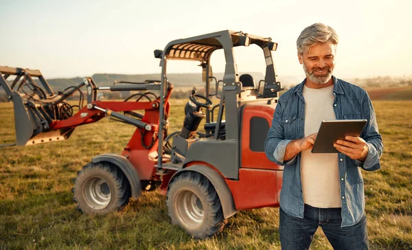 Mature handsome male farmer standing near a tractor with a tablet in the field on a hot day and working on the farm. Agriculture and agriculture concept.
