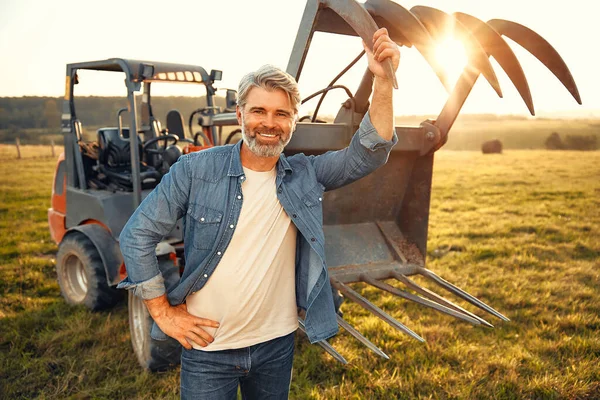 Mature handsome male farmer standing near a tractor on a field on a hot day and working on the farm. Agriculture and agriculture concept.