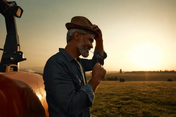 Mature handsome male farmer in a hat standing near a tractor in a field at dusk after a hard day of work. Agriculture and agriculture concept.
