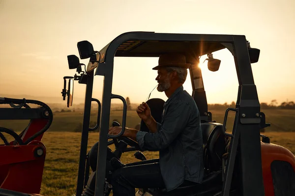 Mature handsome male farmer in a hat sitting in a tractor on a field at dusk after a hard day of work. Agriculture and agriculture concept.