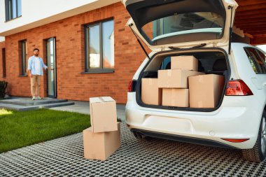 İçinde bir sürü eşya olan açık bir araba bagajı. Arka planda yeni evinin kapısında duran bir adam. Kiralamak ve bir ev almak. Taşınma konsepti.