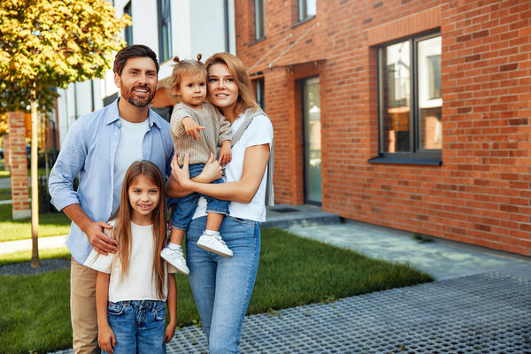 Moving of a young family to a new house. Couple with two kids daughters standing in front of their newly bought house. Renting and buying a home.