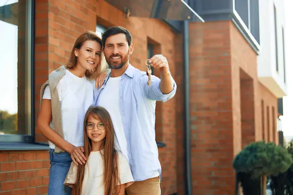 A young family with their daughter standing outside their new house and showing the keys to it. Renting and buying a home. Moving concept.
