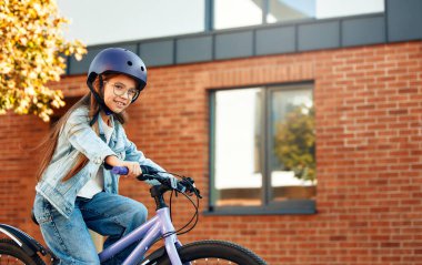 Çocuk dışarıda garaj yolunda bisiklet sürmeyi öğreniyor. Şehirdeki asfalt yolda bisiklet süren küçük bir kız koruyucu giysi olarak kask takıyor..