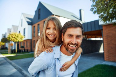 Babası sırtında kızıyla yeni evinin önünde dikilmiş ve seviniyor. Emlak kiralamak ve satın almak. Taşınma konsepti.