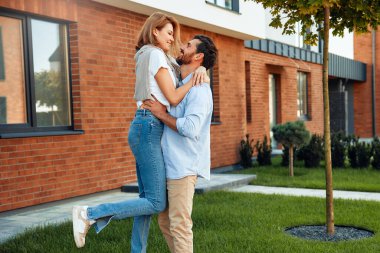 Genç, mutlu, evli bir çift yeni bir evin yanında duruyor ve sarılıyorlar. Emlak kiralamak ve satın almak. Taşınma konsepti.