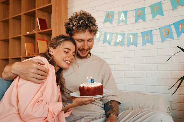 若い結婚したカップルは居心地の良い家のリビングルームに座っている間ケーキにろうそくを吹き飛ばすことによって彼らの誕生日を祝います — ストック写真