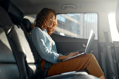 Young business woman with a modern laptop sitting in the back seat of luxury car. Successful startup, distance working, female boss concept.