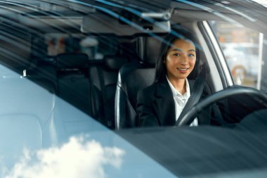 Siyah takım elbiseli, gülümseyen güzel Asyalı genç iş kadını yeni arabasını kullanıyor. Bir araba alıp kiralamak. Seyahat, turizm, eğlence..