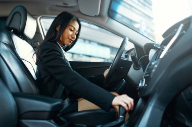 Siyah takım elbiseli, gülümseyen güzel Asyalı genç iş kadını yeni arabasını kullanıyor. Bir araba alıp kiralamak. Seyahat, turizm, eğlence..