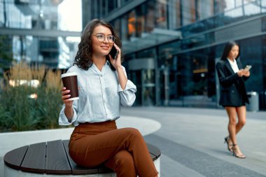 Genç ve güzel bir iş kadını, iş merkezlerinin arka planında otururken hem kahve içip hem de akıllı telefon kullanıyor..