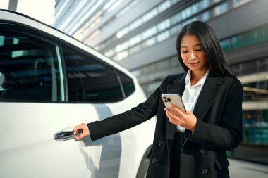 Siyah takım elbiseli güzel, genç Asyalı iş kadını yeni arabasının yanında duruyor ve akıllı telefon kullanıyor. Bir araba alıp kiralamak. Seyahat, turizm, eğlence..