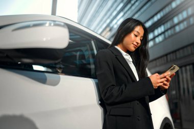 Siyah takım elbiseli güzel, genç Asyalı iş kadını yeni arabasının yanında duruyor ve akıllı telefon kullanıyor. Bir araba alıp kiralamak. Seyahat, turizm, eğlence..