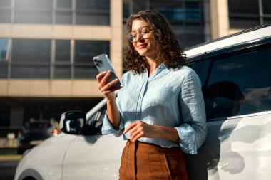Güzel, genç, mutlu, beyaz bir iş kadını yeni arabasının yanında duruyor ve akıllı telefon kullanıyor. Bir araba alıp kiralamak. Seyahat, turizm, eğlence..