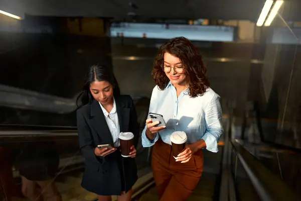 A team of successful business women of different races discuss strategy, drink coffee and use the phone on their way to the office. Diverse women going up the escalator.