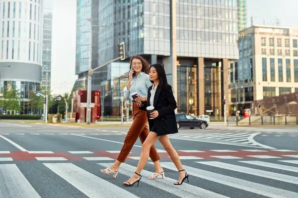 A team of successful business women of different races discuss strategy, drink coffee and use the phone on their way to the office. Diverse women crossing the road at a pedestrian crossing.