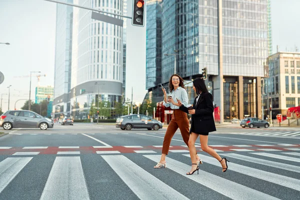 A team of successful business women of different races discuss strategy, drink coffee and use the phone on their way to the office. Diverse women crossing the road at a pedestrian crossing.