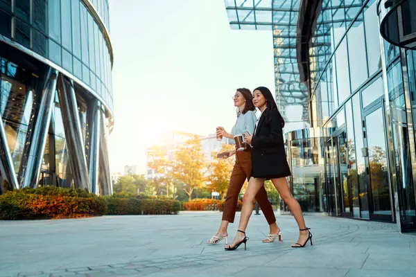Team of multiracial successful business women on their way to the office talking about strategy and drinking coffee. Diverse women walking outside with buildings in the background. Teamwork.