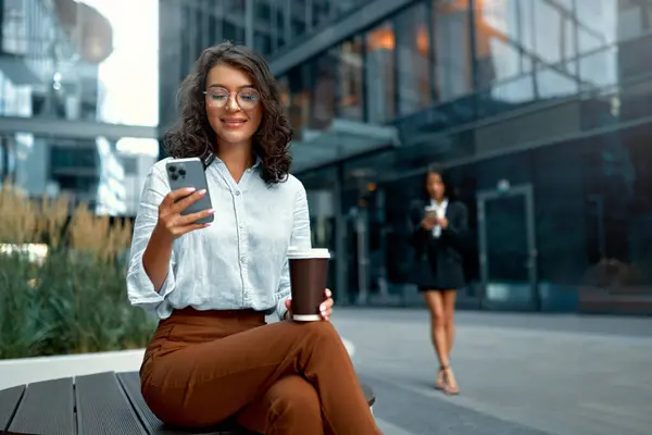A young beautiful business woman drinks coffee and uses a smartphone while sitting on the street against the backdrop of business centers.