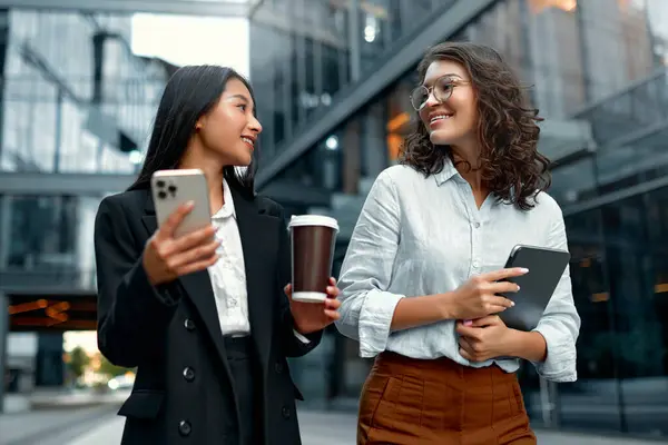 Team of multiracial successful business women on their way to the office talking about strategy, drinking coffee and using phone. Diverse women walking outside with buildings in the background.