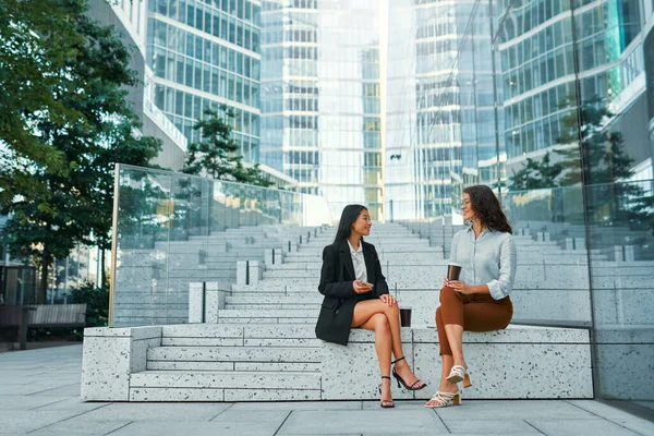 A team of successful business women of different races sitting on the steps discussing strategy, drinking coffee and using the phone. Women walk along the street against the backdrop of buildings.