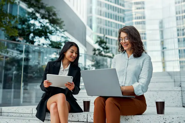 A team of successful business women of different races sitting on the steps with a laptop and tablet, discussing strategy, drinking coffee against the backdrop of business centers.