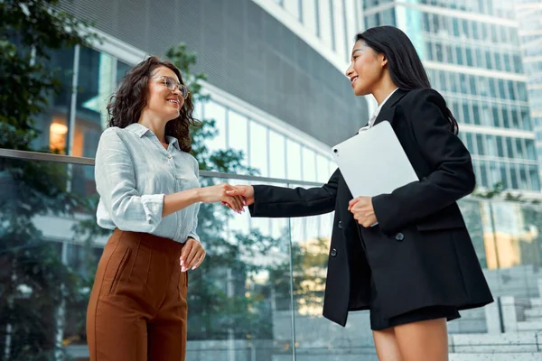 Two business women in business clothes with a tablet shaking hands while standing against the backdrop of business centers.