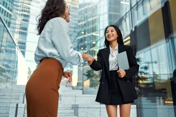 Two business women in business clothes with a tablet shaking hands while standing against the backdrop of business centers.