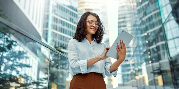 A beautiful business woman in business clothes and glasses holding a tablet while standing on the steps against the backdrop of business centers.