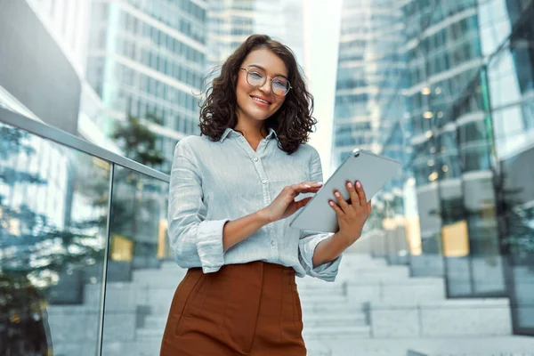 A beautiful business woman in business clothes and glasses holding a tablet while standing on the steps against the backdrop of business centers.