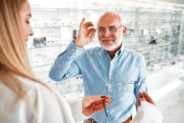 Beyaz önlüklü kadın bir göz doktoru profesyonel bir mağazada yetişkin bir erkeğe optik gözlük gösteriyor. Görüş düzeltme ve göz doktorluğu.