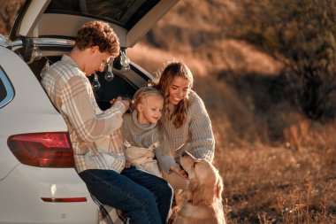 Genç ve mutlu bir aile piknikte sevgili küçük kızları ve köpeğiyle birlikte bir arabanın arkasında oturuyor ve hafta sonu birlikte eğleniyorlar..