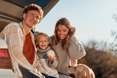 Genç ve mutlu bir aile piknikte sevgili küçük kızları ve köpeğiyle birlikte bir arabanın arkasında oturuyor ve hafta sonu birlikte eğleniyorlar..