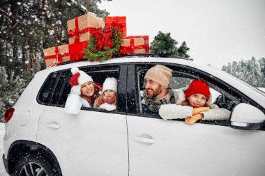 Mutlu Noeller ve mutlu yıllar. Kutular dolusu hediyelerle dolu bir arabada oturan mutlu bir aile ve karlı havada bir ormanın arka planında bir Noel ağacı..