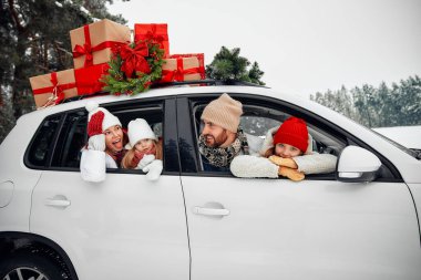 Mutlu Noeller ve mutlu yıllar. Kutular dolusu hediyelerle dolu bir arabada oturan mutlu bir aile ve karlı havada bir ormanın arka planında bir Noel ağacı..
