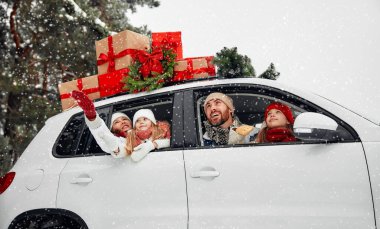 Mutlu Noeller ve mutlu yıllar. Kutular dolusu hediyelerle dolu bir arabada oturan mutlu bir aile ve karlı havada bir ormanın arka planında bir Noel ağacı..