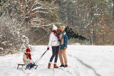 Mutlu Noeller ve mutlu yıllar. Mutlu bir aile, kızakla bir çocuğa biner ve tatil hazırlıkları için karlı bir patika boyunca ormandan bir Noel ağacı taşır..