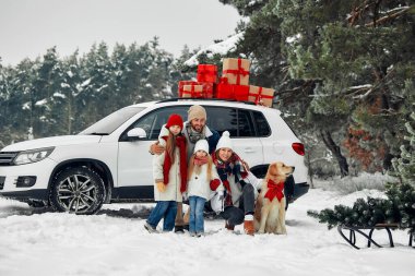 Mutlu Noeller ve mutlu yıllar. Çocuklu bir aile ve hediyelerle dolu bir arabanın yanında duran bir köpek, kızakta duran bir Noel ağacı, karlı bir havada bir ormanın zemininde duran.