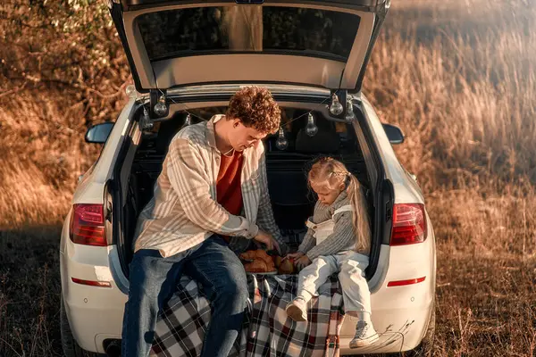 A young happy father on a picnic with his beloved little daughter sitting in the back of a car on a blanket on a weekend having fun together.