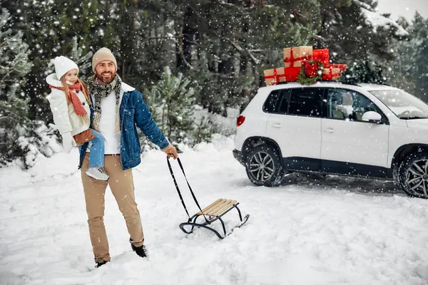 圣诞快乐 新年快乐 一个快乐的父亲 带着孩子和雪橇 站在雪天森林的背景下 满载着礼物的汽车旁边 他走过去捡起一棵圣诞树 — 图库照片
