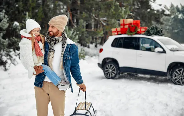 Mutlu Noeller ve yeni yıl. Mutlu çocuklu bir baba ve hediye dolu bir arabanın yanında duran bir kızak. Karlı bir havada bir ormanın arka planında bir Noel ağacı almaya gider..